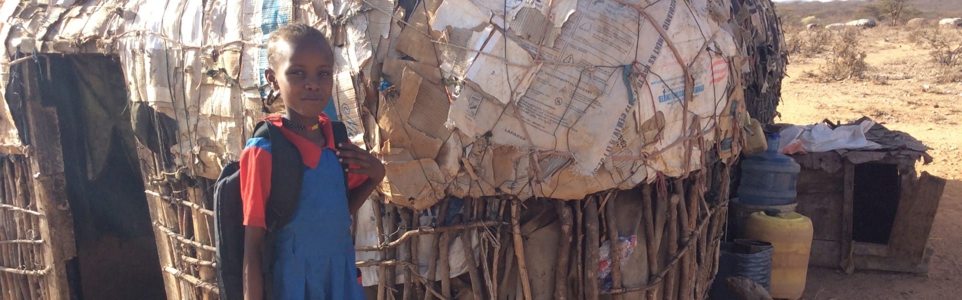 A girl with a backpack outdoors in front of a house in Africa