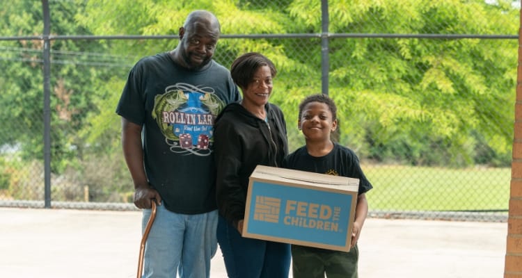 A family standing together outdoors with a feed the children box