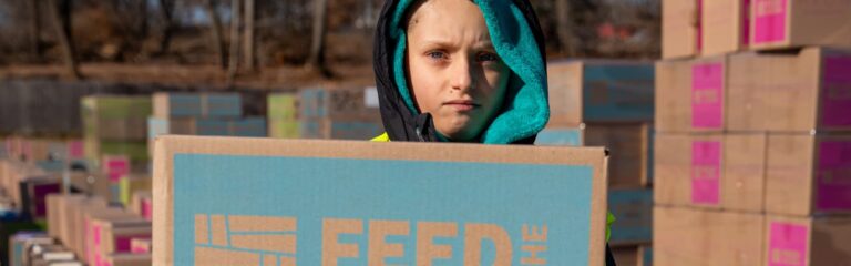 A child frowning with a hood on while standing in front of a box at an event