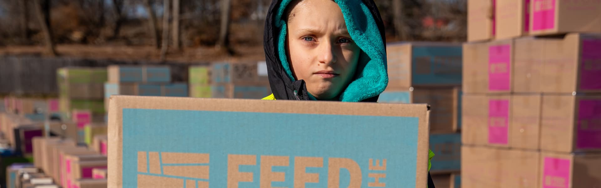 A child frowning with a hood on while standing in front of a box at an event