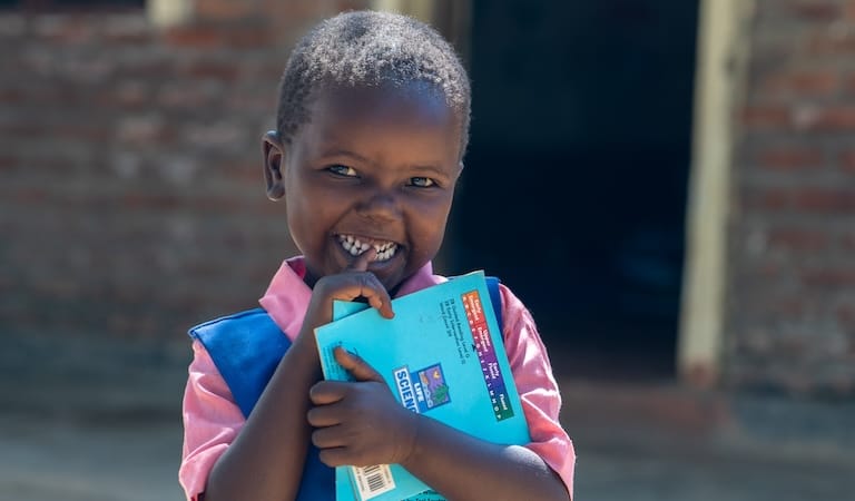 A child standing outdoors holding a book and smiling