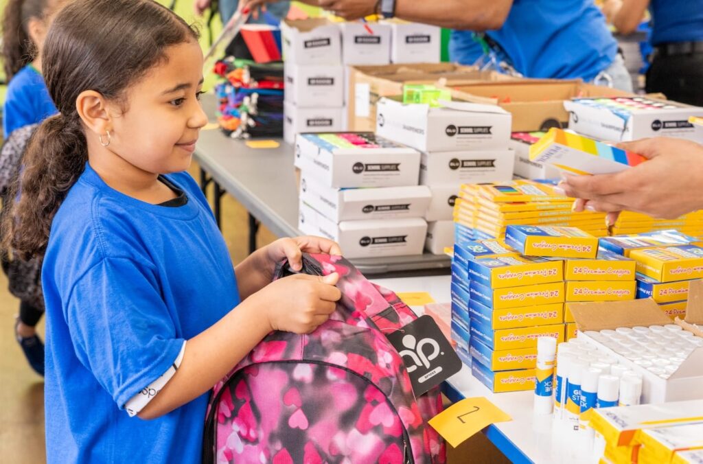 A girl holding a backpack and putting school supplies in it.