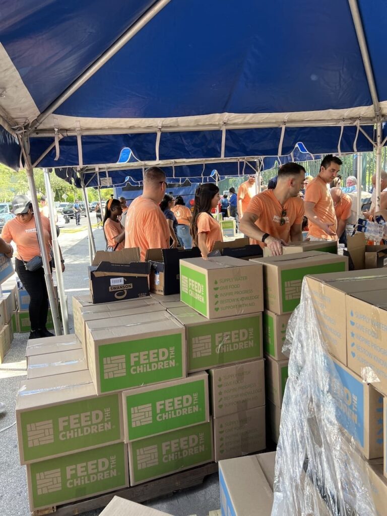 A group of volunteers handing out boxes under a tent with stacks of boxes.