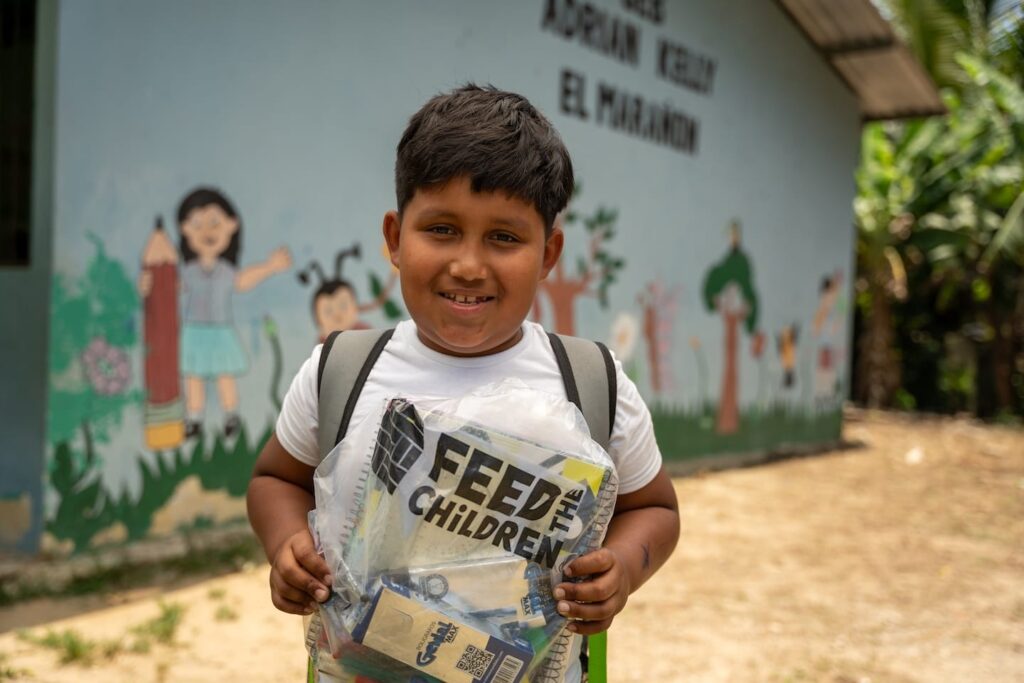 A child standing with a Feed the Children bag outside of a school building.