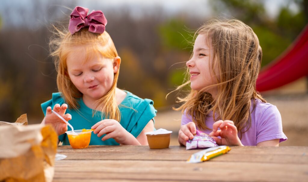Two girls eating fruit cups at a table outdoors.