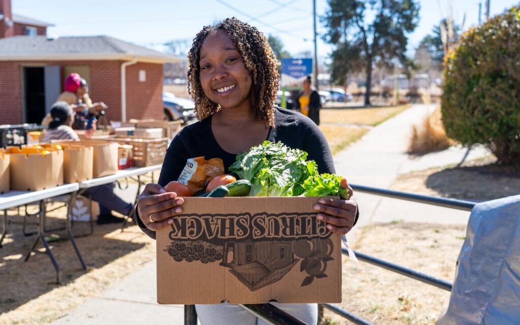 A woman holding a box full of produce at an outdoor event.
