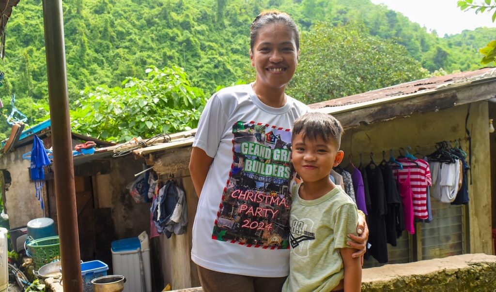 Jeecob with his mother in front of clothes hanging outdoors.