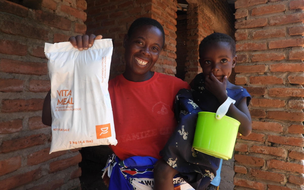 A woman holding a child with a green bucket and a bag of VitaMeal.