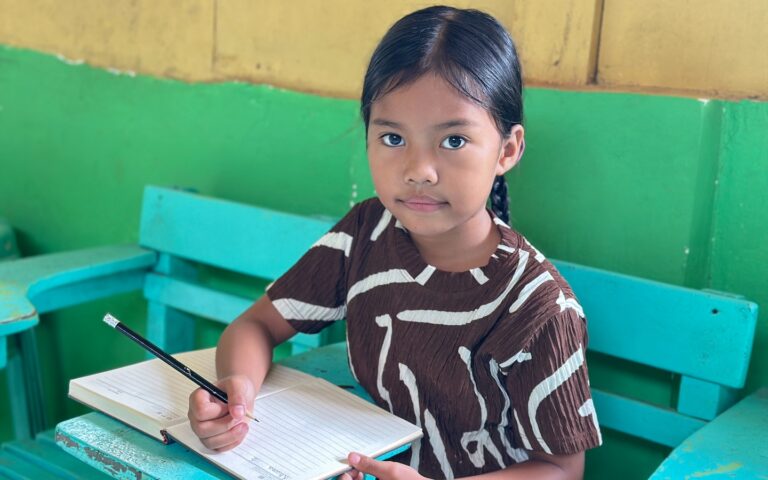 Khianna holding a pencil and sitting at a school desk doing school work.